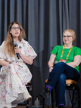 Two panelists on stage at a conference, one speaking into a microphone